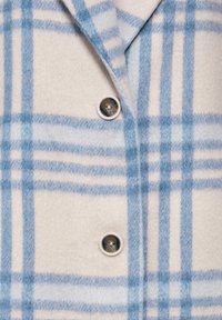 Sininen ja kermanvärinen ruudullinen paitajakku, joka on valmistettu pehmeästä kankaasta. Siinä on viistot kaulukset ja kaksi nappia, ja se sisältää fleecepintaista tekstiiliä.