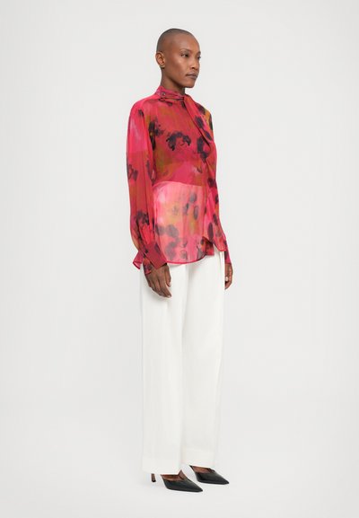 Transparante roze blouse met bloemenpatroon en hoge hals; gecombineerd met witte wijde broek en zwarte puntige hakken.