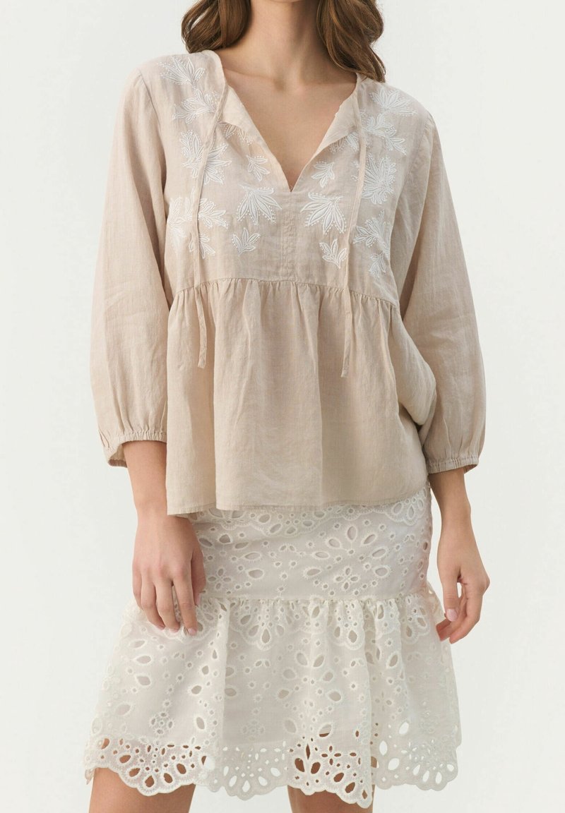 Lichtbeige linnen blouse met witte bloemenborduursels, V-hals en opgezoomde mouwen. Gecombineerd met een witte eyelet kanten rok met een geruffelde zoom.