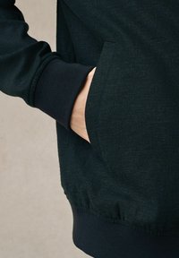 Ciemnozielona bluza z teksturowanym wykończeniem, z boczną kieszenią i ściągaczami przy nadgarstkach. Materiał jest miękki i ma swobodny styl.