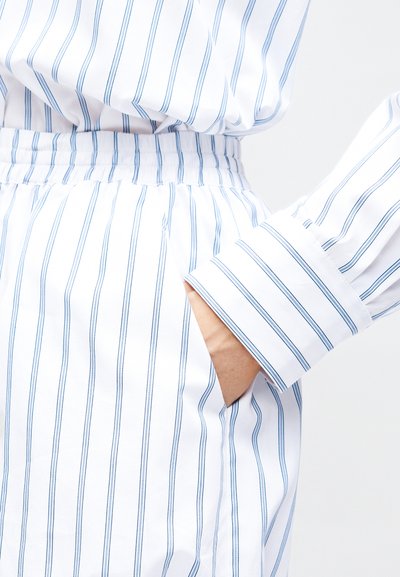 Χέρι στην τσέπη λευκού παντελονιού με μπλε κάθετες ρίγες, συνδυασμένο με μπλούζα μακρυμάνικη με τις ίδιες ρίγες και μανσέτα.