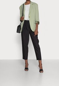 Light green blazer with rolled sleeves, white top, black tapered pants, and black slide sandals. Black crossbody bag hangs from the shoulder.