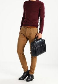 Zwarte lederen aktetas met zilverkleurige hardware, rechthoekige vorm, gedragen door een persoon in een bordeauxrode trui en kaki broek.