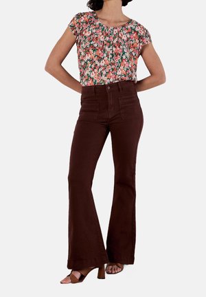 Femme portant un chemisier à manches courtes fleuri, un pantalon flare taille haute marron foncé et des sandales à talons marron, debout les mains dans le dos.