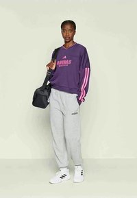 Γυναίκα στέκεται φορώντας μωβ φούτερ Adidas με ροζ ρίγες, γκρι φόρμες Adidas, λευκά αθλητικά παπούτσια και κρατώντας μια μαύρη τσάντα στον ώμο.