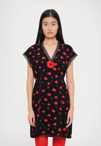 Zwarte jurk met een rood bloemenpatroon, kantafwerking bij de halslijn en een rode bloemenaccent in het midden, gecombineerd met rode panty’s.