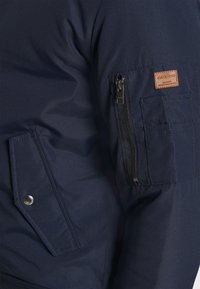 Giacca navy con una tasca con zip sul braccio superiore, dettagli cuciti e un'etichetta logo marrone sulla manica. Tessuto di texture liscia.