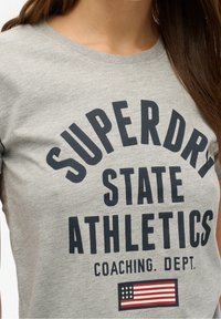 Camiseta de algodón gris con texto negro en negrita "SUPERDRY STATE ATHLETICS COACHING. DEPT." y un pequeño gráfico de la bandera americana.