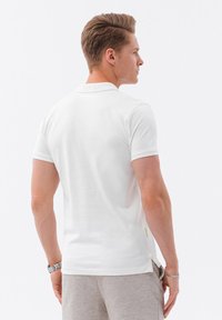 Bílá polo košile vyrobená z bavlny, s klasickým límečkem, krátkými rukávy, bočními rozparky a jemnou značkovou cedulkou na dolním lemu.