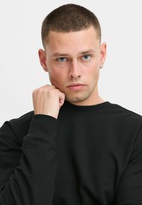 Sudadera negra con cuello redondo, fabricada con una tela suave. El modelo posa con una mano en la barbilla, mostrando su cabello corto y ojos azules.