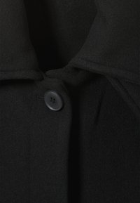 Zwarte jas met een gestructureerde stof, notch kraag en een enkele zwarte knoopdetail aan de voorkant. Geen zichtbare patronen of accenten.