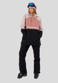 Vrouw in een roze en zwarte snowboardjack, zwarte sneeuwbroek, zwarte skibotten, een roze beanie en blauwe skibril op haar voorhoofd.