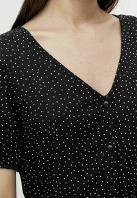Czarna bluzka z dekoltem w serek, krótkimi rękawami i białymi kropkami. Posiada guzikowe zapięcie z czarnymi guzikami oraz luźny krój.