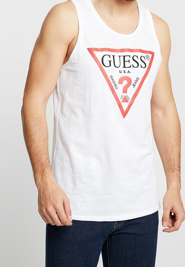 Débardeur blanc en coton avec un logo triangulaire rouge affichant les mots « GUESS » et « U.S.A. », associé à un jean en denim bleu. Pas de motifs ou d'accents remarquables.