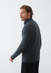Un pull en tricot gris avec un col montant, un motif texturé et des manches longues, porté avec un pantalon sombre, vu de côté.