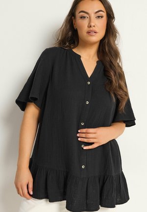 Kvinde iført løs sort skjorte med knapper, flæset kant og korte ærmer, poserer med den ene hånd på hoften mod en ensfarvet baggrund.