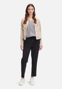 Veste courte beige clair avec deux poches frontales sur un haut gris texturé à col en V, assortie à un pantalon noir fuselé et des baskets blanches.