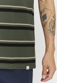 Camiseta de rayas verdes con rayas horizontales en negro y beige, material de algodón, y una pequeña etiqueta blanca en el dobladillo. Mano visible con tatuajes.