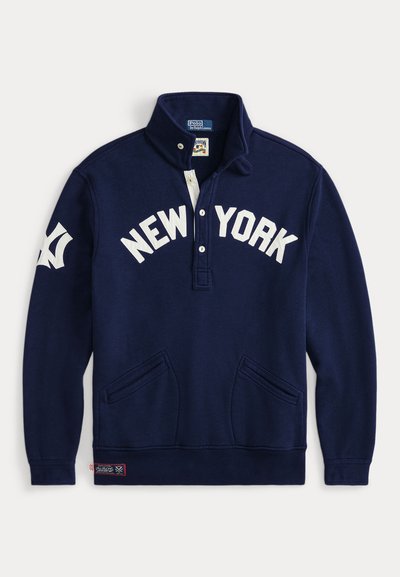 Marinblå tröja med vit text "NEW YORK", två framfickor, halv-knäppkrage och logotyp på ärmen. Mjuk, texturerad tyg.