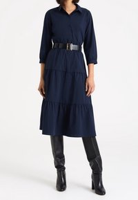 Vestido azul marino de capas con botones en la parte delantera y cuello, ceñido a la cintura con un cinturón negro, combinado con botas negras hasta la rodilla.