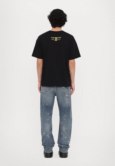 T-shirt en coton noir à coupe ample, arborant "Craft With Pride" en jaune dans le dos. Porté avec un jean bleu déchiré et des chaussures noires.