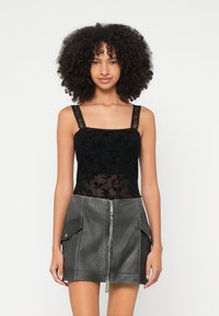 Mulher com cabelo encaracolado, vestindo uma camisola preta de renda e uma minissaia cinzenta de imitação de couro, com fecho éclair frontal e bolsos laterais.