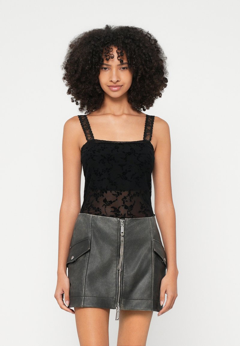 Mulher com cabelo encaracolado, vestindo uma camisola preta de renda e uma minissaia cinzenta de imitação de couro, com fecho éclair frontal e bolsos laterais.