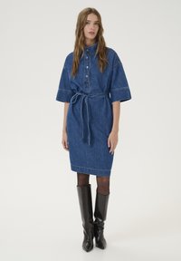 Denim-paitamekko, jossa on kauluskanta, lyhyet hihat ja kietaistu vyö vyötäröllä. Tumansinisestä kankaasta, jossa on sileä pinta ja näkyvät ompeleet.
