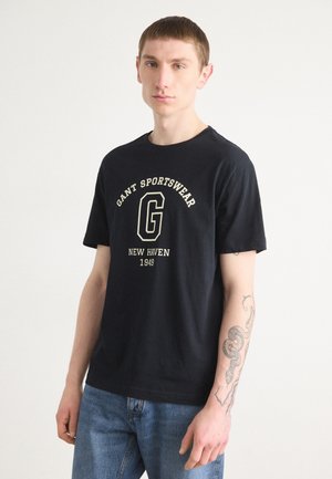 T-shirt in cotone nero con maniche corte, caratterizzato da una stampa grafica bianca e dorata con scritto "GANT SPORTSWEAR NEW HAVEN 1949." Texture liscia.
