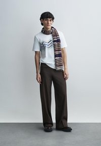 Λευκό μπλουζάκι με ζιγκ ζαγκ μοτίβο σε μπλε και πράσινο, συνδυασμένο με καφέ παντελόνια φαρδιάς γραμμής και ριγέ φουλάρι σε μπορντό και κρεμ.