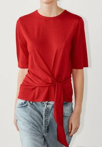 Top rosso a maniche corte realizzato in tessuto morbido, con un dettaglio annodato in vita e un collo rotondo. Indossato con jeans in denim blu chiaro.