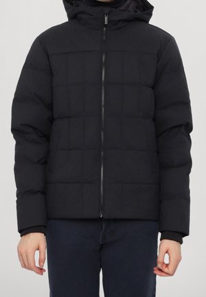 Mann trägt eine schwarze, gesteppte Steppjacke mit Frontreißverschluss und Kapuze, kombiniert mit dunkelblauer Hose, stehend vor einem schlichten Hintergrund.