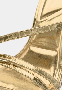 Auksinės spalvos metalinės aukštakulnės basutės su daugybe plonų dirželių, blizgiu paviršiumi ir lygia padu. Matomos tekstūruotos detalės ant dirželių.