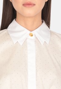 Blusa trasparente color beige chiaro con colletto bianco, chiusura a bottoni frontale e borchie in argento incorporate per texture e dettaglio.