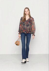Blusa solta de várias cores com mangas longas e soltas, combinada com jeans azuis em boca de sino e botins bege. O modelo segura uma pequena mala bege.