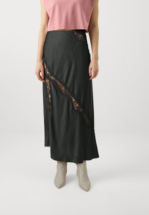 Femme portant une jupe midi en satin noir avec des panneaux de dentelle en diagonale, des bottines pointues couleur pâle, et un haut court rose.