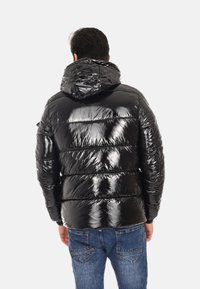 Giacca a vento nera con una texture lucida e trapuntata e cappuccio. Presenta cuciture orizzontali e una vestibilità comoda. Jeans visibili in basso.