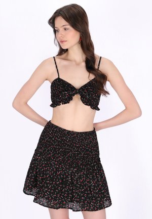 Młoda kobieta o falowanych brązowych włosach, ubrana w czarny kwiatowy krótki top na cienkich ramiączkach oraz dopasowaną, rozkloszowaną spódnicę, z rękami na biodrach, patrzy w lewo.