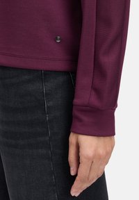 Bordowy sweter z długim rękawem, o miękkiej fakturze i zaokrąglonym dole. Posiada mały czarny guzik przy dolnym brzegu. Noszony z ciemnymi dżinsami.