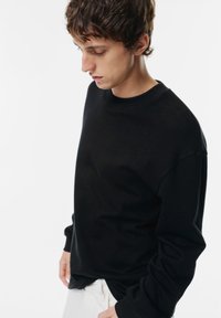 Maglione nero a maniche lunghe con scollatura rotonda, realizzato in tessuto testurizzato, leggermente oversize su pantaloni bianchi.