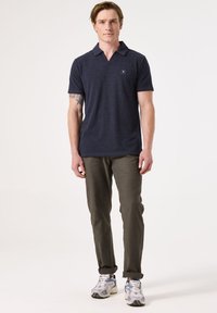 Marineblauwe poloshirt met een kraag, korte mouwen en een vierkant logo op de borst. Olijfgroene broek met een gladde textuur en opgerolde manchetten. Grijze sneakers.