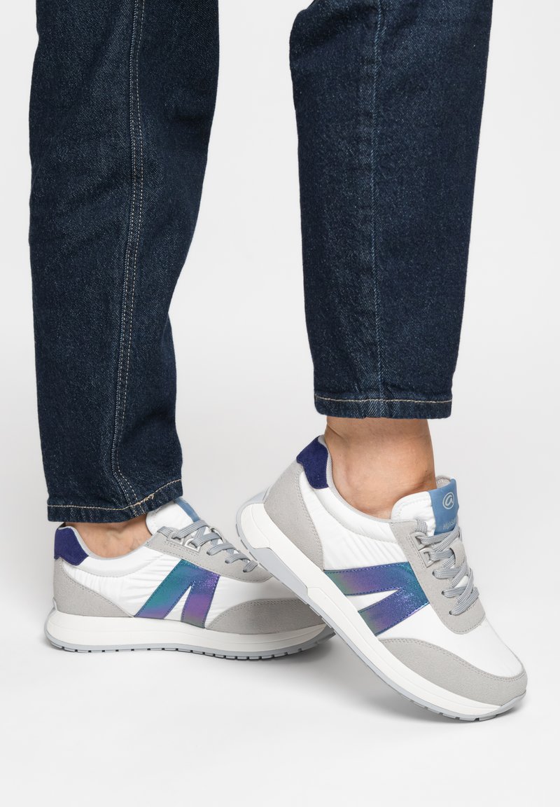 Personne portant un jean bleu et des baskets blanches avec des accents gris et bleu violet irisé sur un fond blanc.