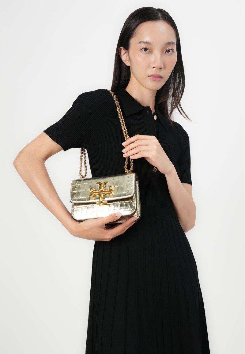 Metallisch goldene Handtasche mit Krokodilprägung, Verschluss und Kettenriemen, die in der rechten Hand gehalten wird, vor einem einfarbigen Hintergrund.