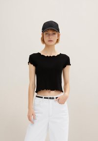 Svart ribbad kort topp med volanger i kanten, matchad med högmidjade vita jeans och en svart keps, med en elegant design och minimal hårdvara.