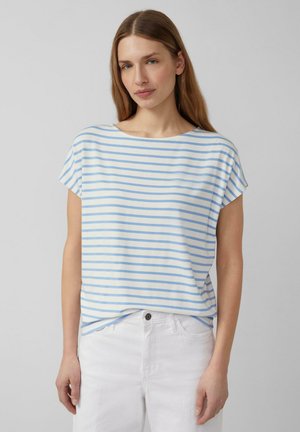 Vrouw met steil lichtbruin haar, gekleed in een wit- en lichtblauw gestreept shirt met korte mouwen en een witte broek, tegen een effen achtergrond.