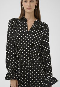 Schwarzes Langarmkleid mit cremefarbenen Polka-Dots, V-Ausschnitt und Taillengürtel. Mit Rüschensäumen an den Ärmeln und einer glatten Stoffstruktur.