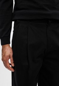 Linkerhand rustend naast zwarte plissébroek en zwart langslavend shirt van een persoon, waarbij de stoftextuur en naadtoeslagen zichtbaar zijn.