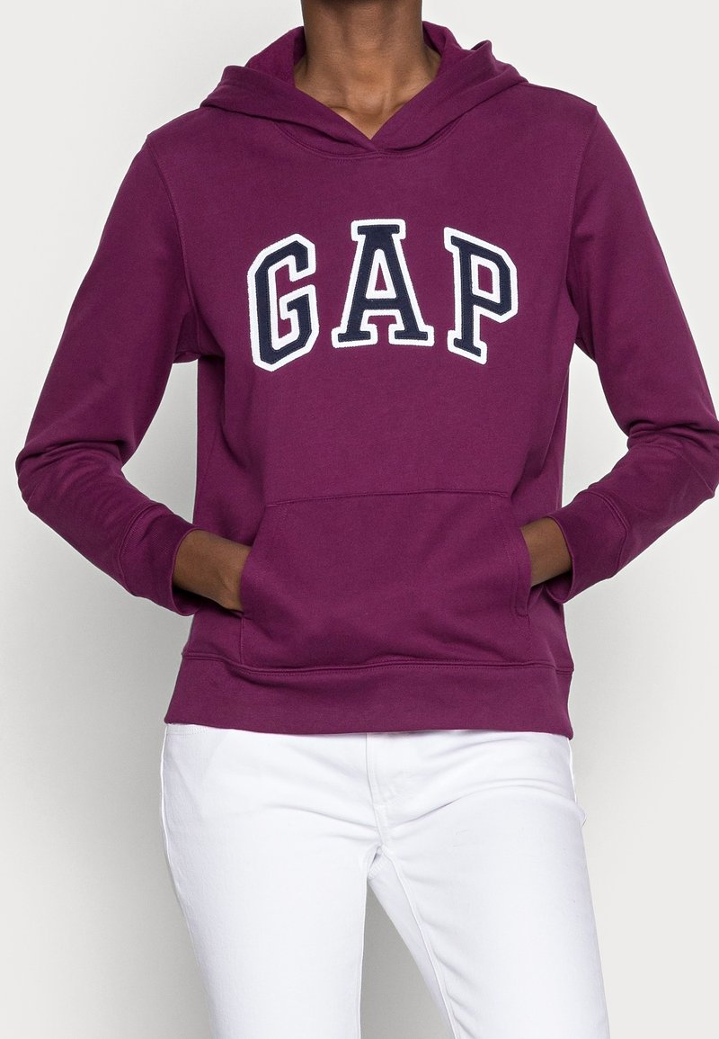 Personne portant un sweat à capuche violet avec le logo "GAP" en grandes lettres blanches et noires, ainsi qu'un pantalon blanc, les mains dans la poche du sweat.