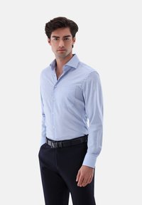 Mann in einem hellblauen karierten Hemd mit dunkler Hose und schwarzem Gürtel, vor einem einfarbigen hellen Hintergrund stehend.