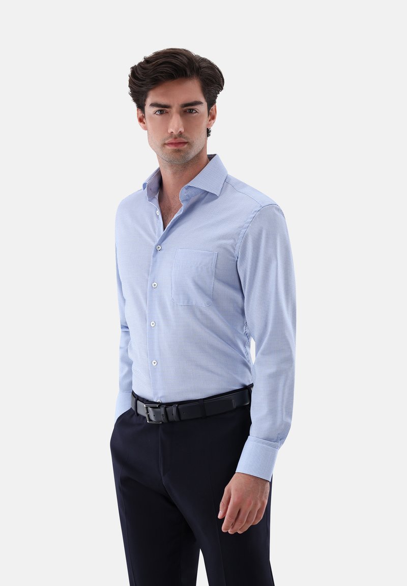Mann in einem hellblauen karierten Hemd mit dunkler Hose und schwarzem Gürtel, vor einem einfarbigen hellen Hintergrund stehend.
